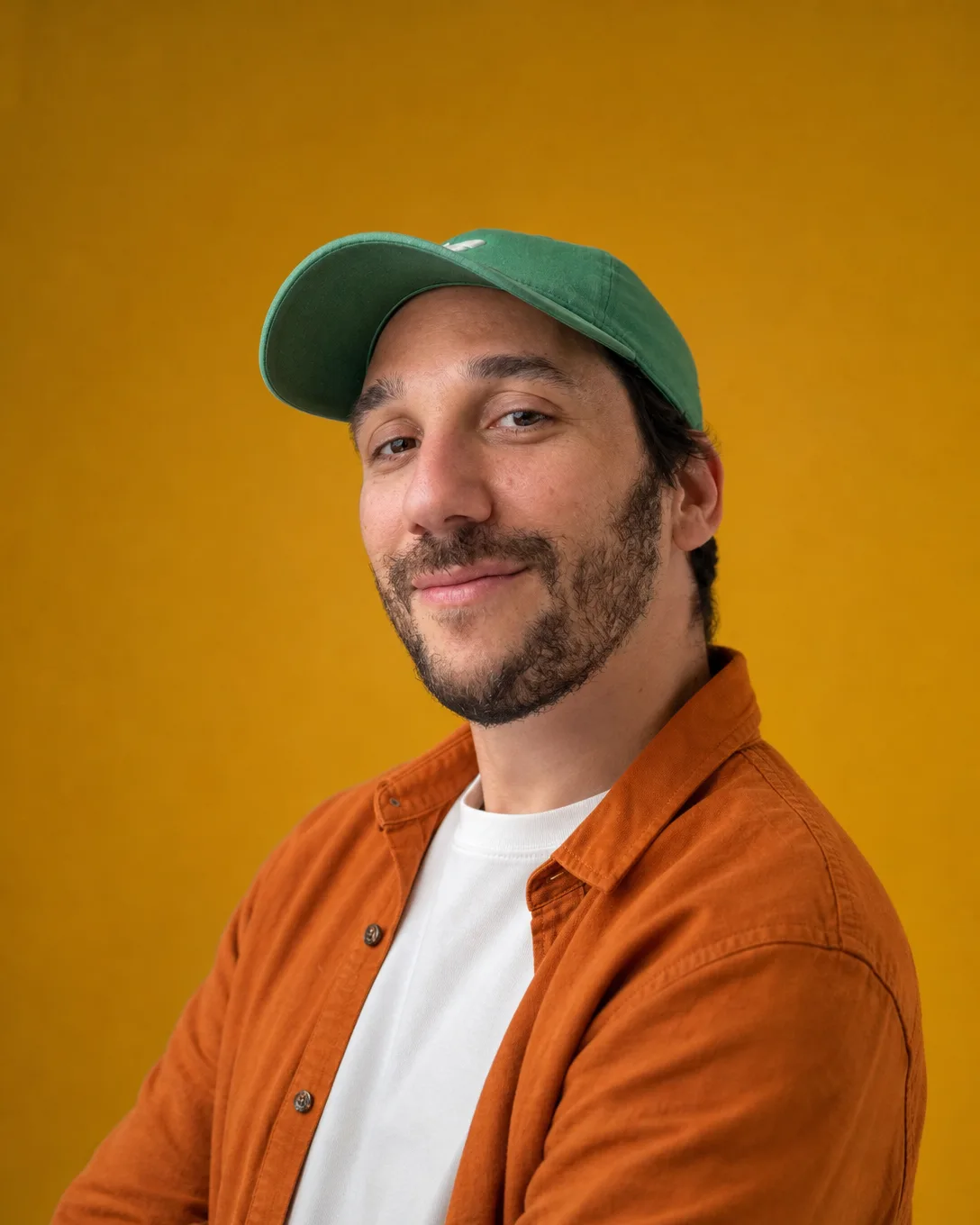 Portrait d'Ismael sur un fond jaune moutarde, surchemise orange sur un t-shirt blanc, casquette verte, sourire