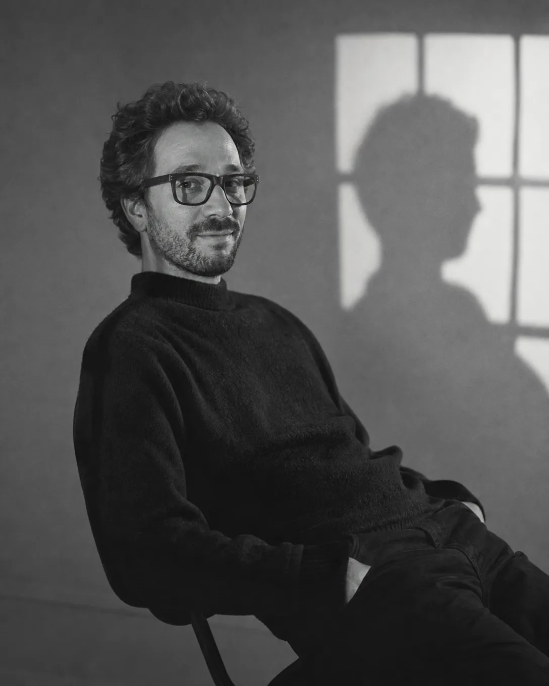 Portrait en noir et blanc d'Ismael assis, lunettes, pull sombre, l'ombre d'une fenêtre sur le mur